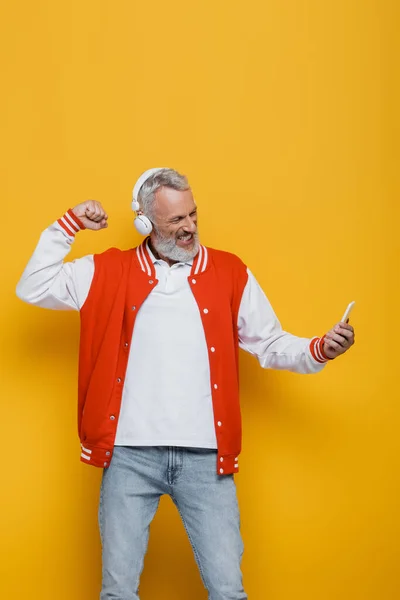 Feliz hombre de mediana edad en auriculares tomando selfie mientras muestra puño apretado en amarillo - foto de stock