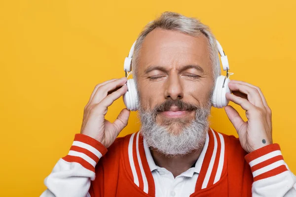 Positive middle aged man in wireless headphones listening music isolated on yellow — Stock Photo