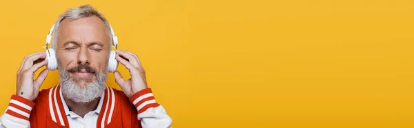 Задоволений чоловік середнього віку в бездротових навушниках прослуховування музики ізольовано на жовтому, банер — стокове фото