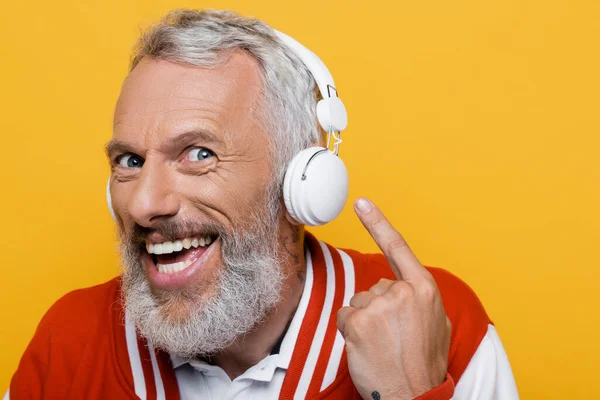 Позитивний чоловік середнього віку вказує на бездротові навушники і слухає музику ізольовано на жовтому — стокове фото
