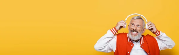 Homem de meia-idade positivo segurando fones de ouvido sem fio isolados em amarelo, banner — Fotografia de Stock