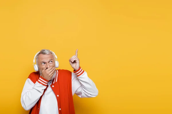 Middle aged man in wireless headphones listening music while covering mouth and pointing up on yellow — Stock Photo