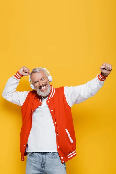 Joyful middle aged man in wireless headphones listening music and dancing on yellow — Stock Photo