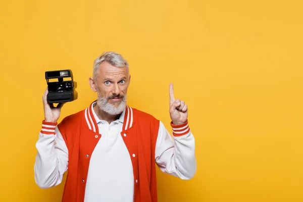 Homme d'âge moyen en veste de bombardier tenant caméra vintage noire et pointant vers le haut isolé sur jaune — Photo de stock