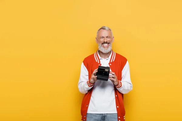 Alegre hombre de mediana edad en chaqueta de bombardero con cámara vintage negro aislado en amarillo - foto de stock