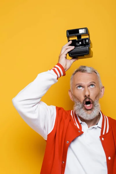 Middle aged man in bomber jacket holding black vintage camera above head isolated on yellow — Stock Photo