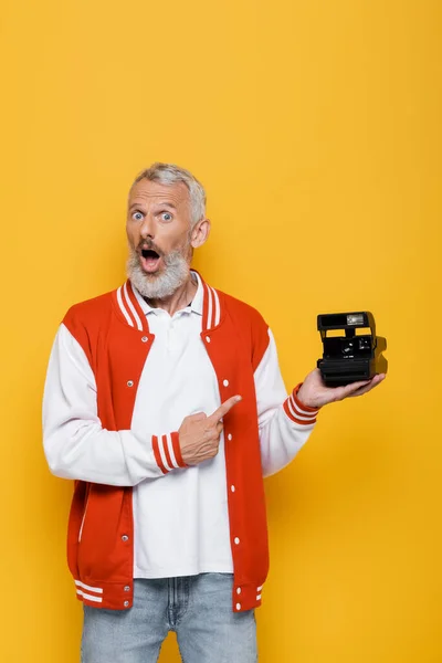 Chocado homem de meia idade em jaqueta bomber apontando para câmera vintage preto isolado no amarelo — Fotografia de Stock