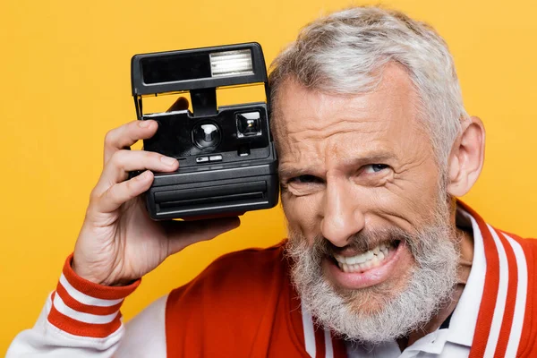Middle aged man in bomber jacket holding black vintage camera and grinning isolated on yellow — Stock Photo