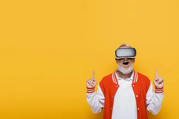 Sorprendido hombre de mediana edad en chaqueta de bombardero y auriculares vr apuntando hacia arriba aislado en amarillo - foto de stock