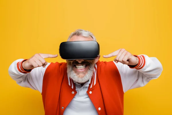 Fröhlicher Mann mittleren Alters in Bomberjacke zeigt auf vr Headset isoliert auf gelb — Stockfoto