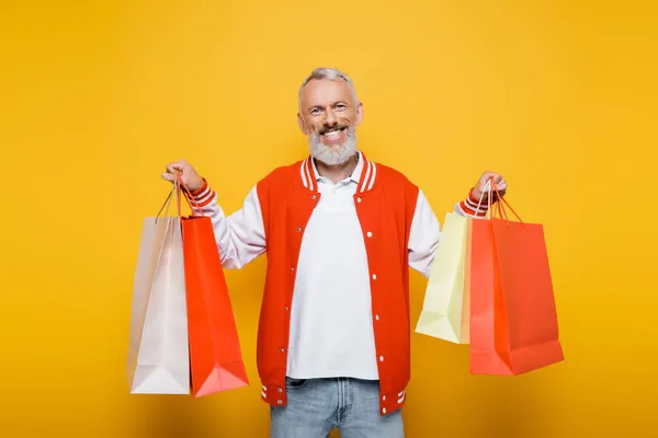 Feliz hombre de mediana edad en chaqueta bombardero sosteniendo bolsas de compras en amarillo - foto de stock