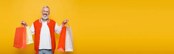 Feliz homem de meia idade em jaqueta bombardeiro segurando sacos de compras isolados em amarelo, banner — Fotografia de Stock