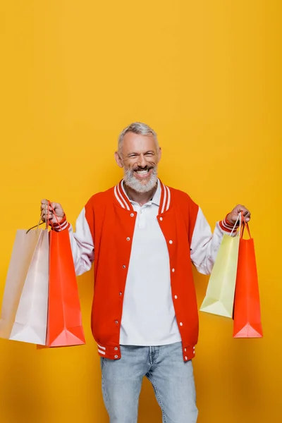 Joyful middle aged man in bomber jacket holding shopping bags on yellow — Stock Photo