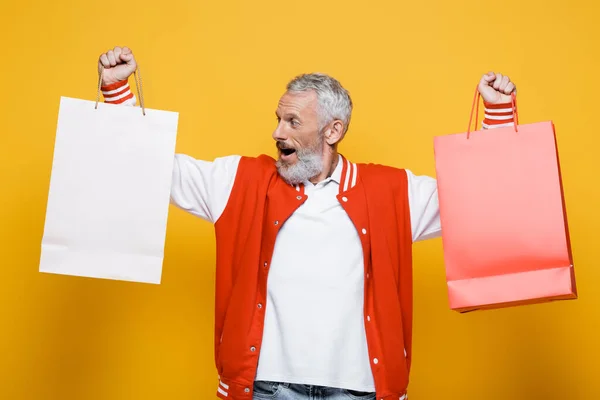 Sorprendido hombre de mediana edad en chaqueta bombardero sosteniendo bolsas de compras en amarillo - foto de stock