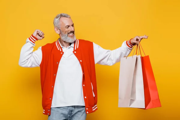 Complacido hombre de mediana edad en chaqueta bombardero sosteniendo bolsas de compras en amarillo - foto de stock