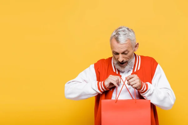 Изумленный мужчина средних лет в куртке подрывника держит сумку с покупками на желтом — стоковое фото