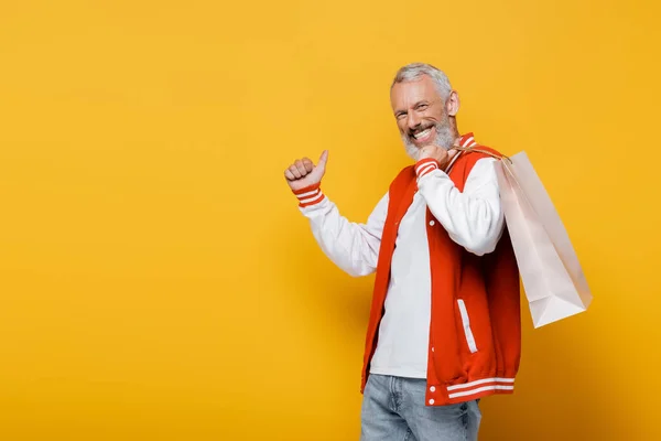 Feliz hombre de mediana edad en chaqueta de bombardero sosteniendo bolsa de compras y mostrando el pulgar hacia arriba en amarillo - foto de stock