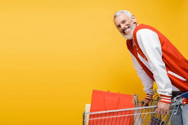 Complacido hombre de mediana edad en chaqueta de bombardero cerca del carro con bolsas de compras aisladas en amarillo - foto de stock