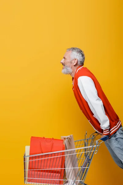 Vista laterale di felice uomo di mezza età in giacca bomber vicino al carrello con borse della spesa su giallo — Foto stock