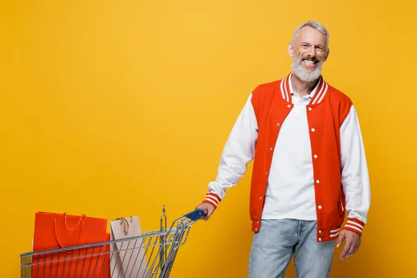 Cheerful middle aged man in bomber jacket standing near cart with shopping bags on yellow — Stock Photo