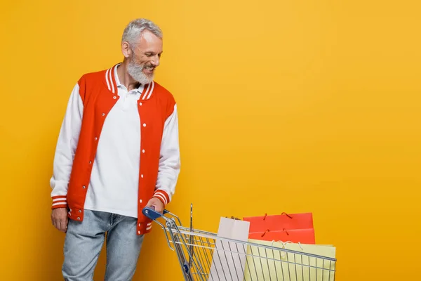 Complacido hombre de mediana edad en chaqueta de bombardero mirando al carrito con bolsas de compras en amarillo - foto de stock