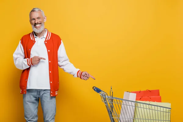 Zufriedener Mann mittleren Alters in Bomberjacke zeigt auf Einkaufswagen mit gelben Einkaufstüten — Stockfoto