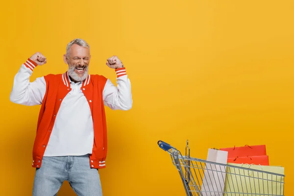 Excitado hombre de mediana edad en chaqueta bombardero regocijo cerca de carro con bolsas de compras en amarillo - foto de stock