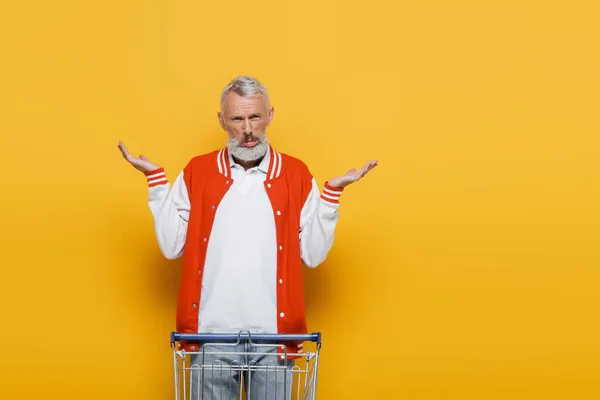 Confused middle aged man in bomber jacket showing shrug gesture near empty shopping cart on yellow — Stock Photo