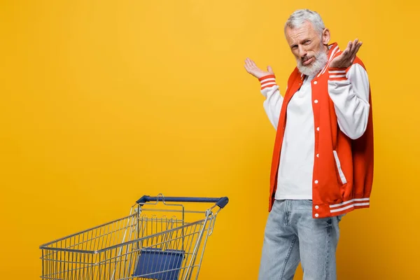 Hombre de mediana edad en chaqueta de bombardero mostrando gesto encogiéndose de hombros cerca del carrito de compras vacío en amarillo - foto de stock