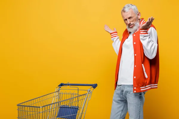 Middle aged man in bomber jacket showing shrug gesture near empty shopping cart on yellow — Stock Photo
