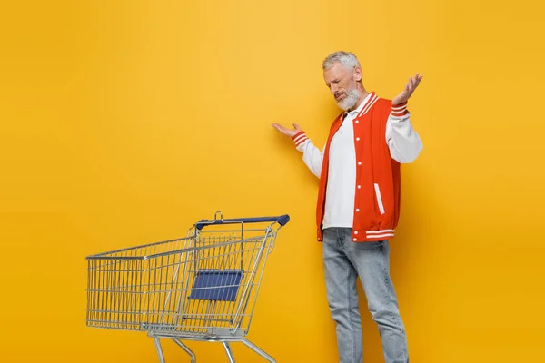 Descontento hombre de mediana edad en chaqueta de bombardero mostrando gesto encogiéndose de hombros cerca de la cesta de la compra vacía en amarillo - foto de stock