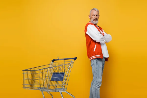 Descontento hombre de mediana edad en chaqueta de bombardero de pie con los brazos cruzados cerca de la cesta de la compra vacía en amarillo - foto de stock