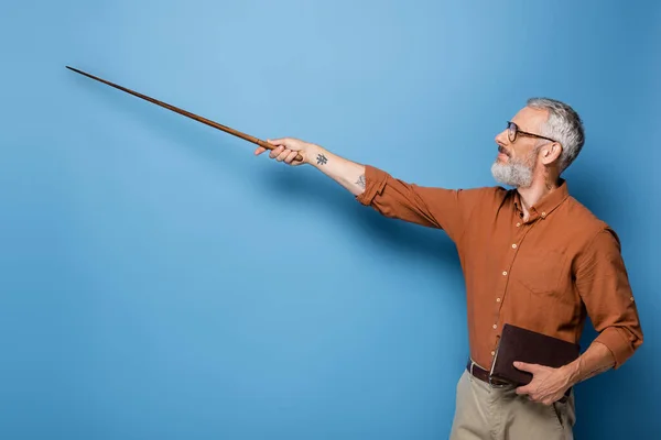 Tatoué enseignant d'âge moyen dans des lunettes tenant pointeur bâton et livre sur bleu — Photo de stock