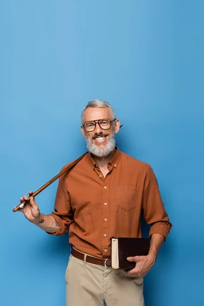 Happy middle aged teacher in glasses holding pointer stick and book on blue — Stock Photo