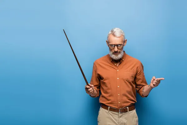 Tattooed middle aged teacher in glasses holding pointer stick and pointing away on blue — Stock Photo