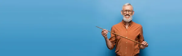 Happy middle aged teacher in glasses holding pointer stick on blue, banner — Stock Photo