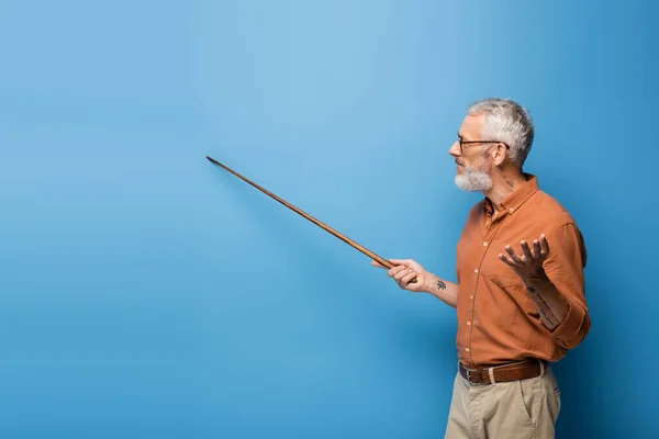 Tattooed middle aged teacher in glasses holding pointer stick and gesturing on blue — Stock Photo