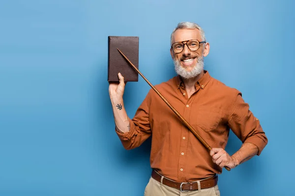 Felice insegnante di mezza età in bicchieri tenendo bastone puntatore vicino libro sul blu — Foto stock