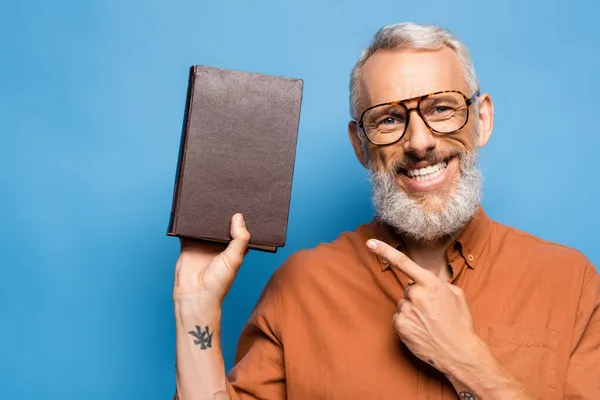 Щасливий вчитель середнього віку в окулярах, вказуючи на книгу на синьому — стокове фото