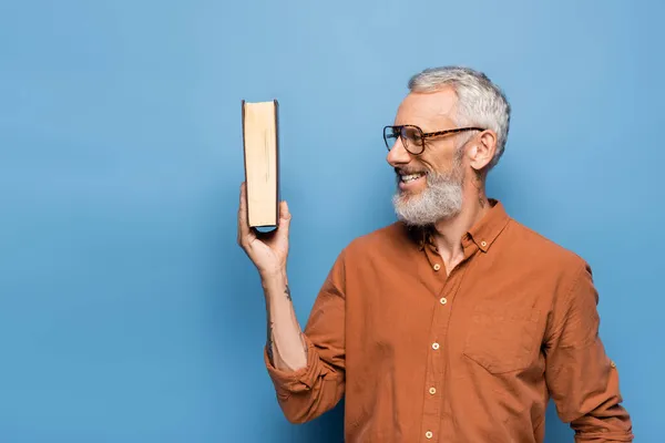 Tattooed middle aged teacher in glasses smiling and holding book on blue — Stock Photo