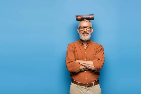 Задоволений вчитель середнього віку в окулярах і книга на голові на синьому — стокове фото