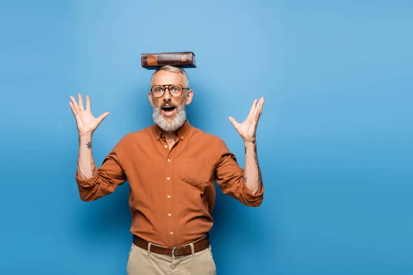 Asombrado maestro de mediana edad en gafas y libro sobre la cabeza gesto en azul - foto de stock