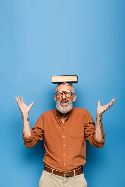 Tatoué moyen âge enseignant dans des lunettes et livre sur la tête gestuelle sur bleu — Photo de stock