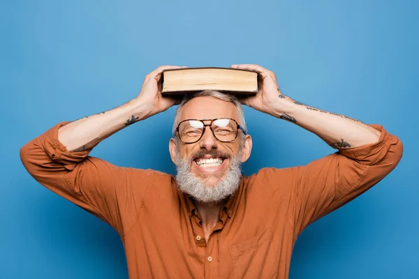 Gioioso insegnante di mezza età in bicchieri tenendo libro sulla testa su blu — Foto stock