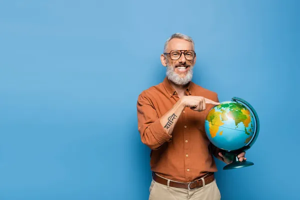Feliz maestro de mediana edad en gafas apuntando con el dedo al globo en azul - foto de stock