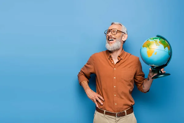Happy middle aged teacher in glasses holding globe and standing with hand on hip on blue — Stock Photo