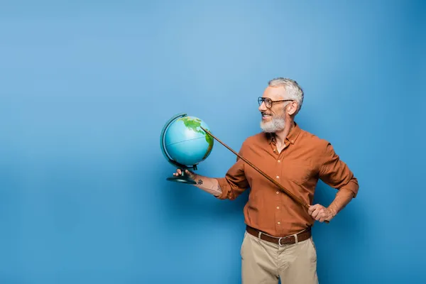 Allegro insegnante di mezza età in occhiali tenendo bastone puntatore vicino al globo su blu — Foto stock