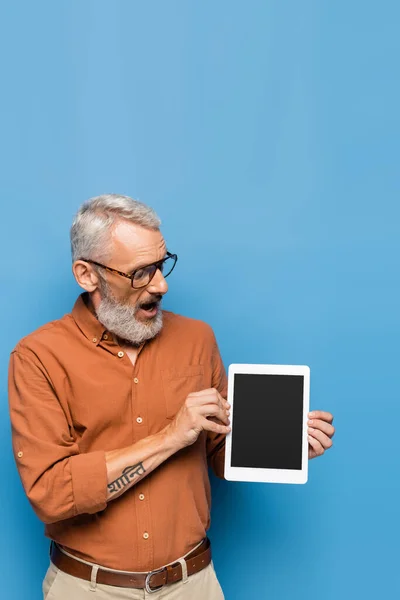 Удивленный мужчина средних лет в очках и рубашке, глядя на цифровой планшет с чистым экраном на синем — стоковое фото