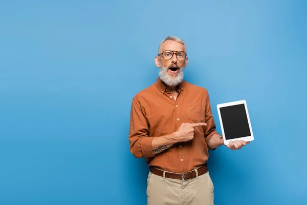 Überraschter Mann mittleren Alters in Brille und Hemd zeigt auf digitales Tablet mit leerem Bildschirm auf blauem Hintergrund — Stockfoto