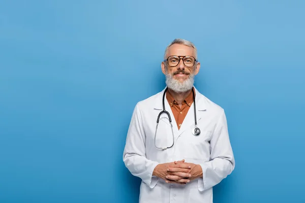 Счастливый врач средних лет в очках, стоящий с сжатыми руками на синем — стоковое фото